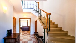 Un escalier avec une main courante en bois et des balustrades en métal, menant à l’étage supérieur. Un banc est placé près de l’escalier, et une porte est visible sur le côté gauche.
