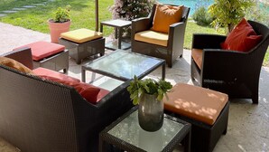 A covered outdoor seating area with wicker furniture, a glass table, and a view of a house and swimming pool. - Superbe Villa Avec Piscine, Idéale Pour Vacances en Famille (ROQUEEFORT LES PINS)