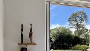 A bedroom with a bed, a window with a view of a pool and greenery, and wall-mounted shelves with a clock and a giraffe figurine. - Superbe Villa Avec Piscine, Idéale Pour Vacances en Famille (ROQUEEFORT LES PINS)