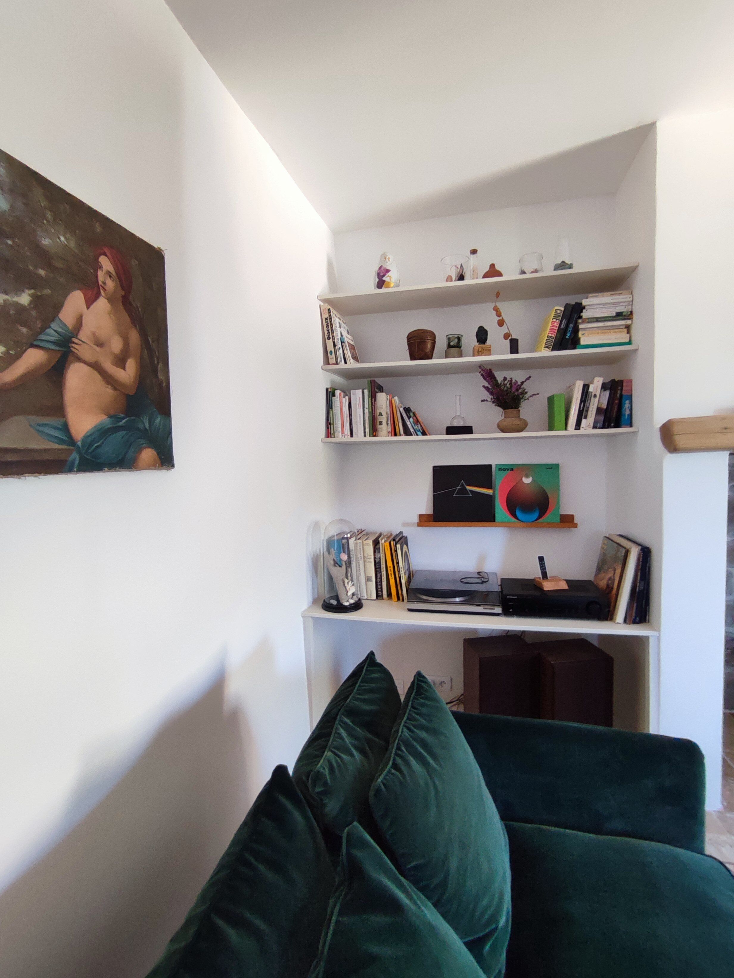 Een woonkamer met een groene bank, een boekenkast vol boeken en decoratieve objecten, en een schilderij aan de muur.