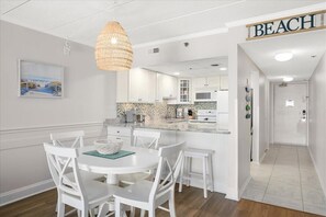 Une cuisine moderne avec des meubles blancs, un coin repas avec des chaises blanches, un luminaire suspendu et un panneau indiquant « PLAGE ».