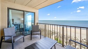 Ein Balkon mit Blick auf den Strand, ein Stuhl, ein Tisch und eine Glastür, die zu einem Zimmer führt.