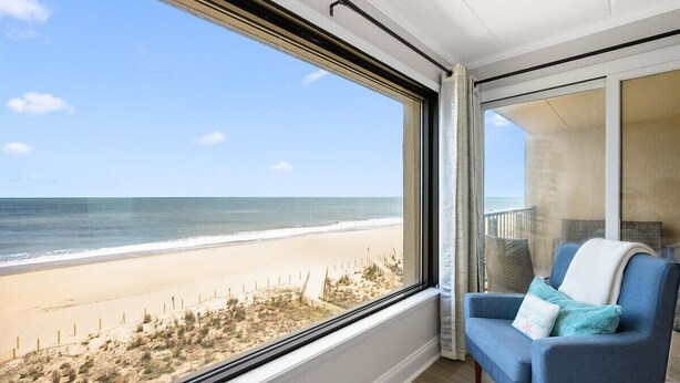 Ein Zimmer mit großem Fenster mit Blick auf einen Strand, ein blauer Sessel und Meerblick.