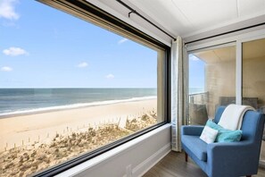 Une pièce dotée d’une grande fenêtre donnant sur une plage, d’un fauteuil bleu et d’une vue sur l’océan.