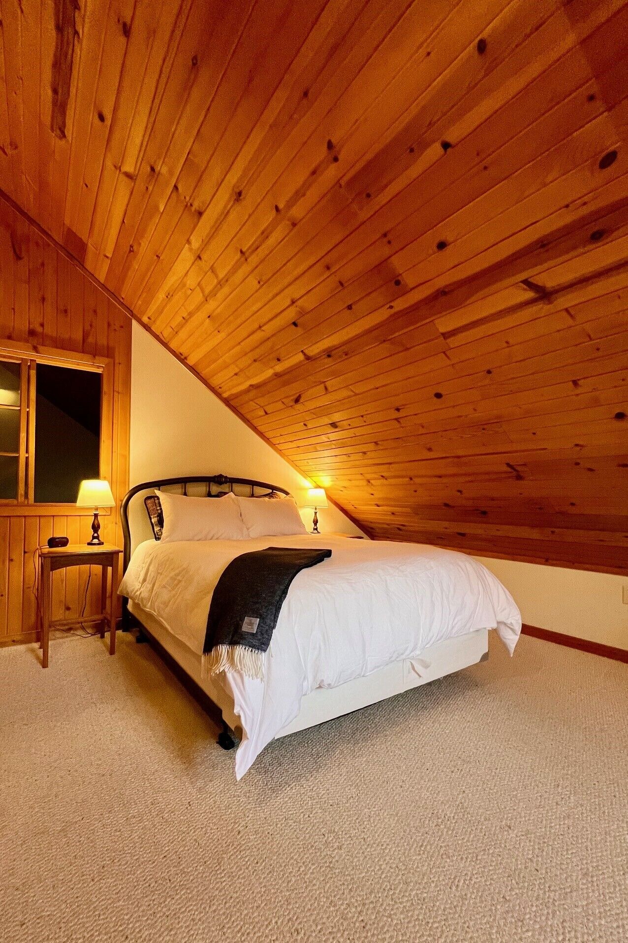 A bedroom with a wooden ceiling, a bed with white bedding, a bedside table with a lamp, and a window with a view.