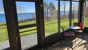 Een kamer met grote ramen met uitzicht op een grasveld en een waterlichaam. Binnen in de kamer staat een tafel en twee stoelen.