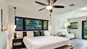 A modern bedroom with a large bed, a ceiling fan, a bench, a desk, and a view of the outdoors. - Windward Escape (Pompano Beach)