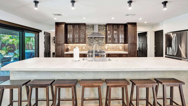A modern kitchen with a large island, dark wooden cabinets, a stone backsplash, and a stainless steel refrigerator. - Windward Escape (Pompano Beach)