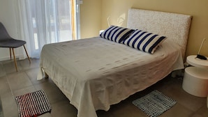 A bedroom with a bed, a chair, a bedside table, and a window with curtains. - Bassin d'Arcachon: couple, quiet, bright, comfortable house with all amenities (Gujan-Mestras)