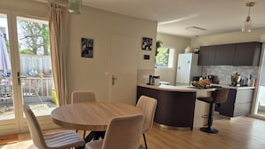 A modern kitchen with a central island, a dining area with a round table and chairs, and a view of the outdoors through a sliding glass door. - Bassin d'Arcachon: couple, quiet, bright, comfortable house with all amenities (Gujan-Mestras)