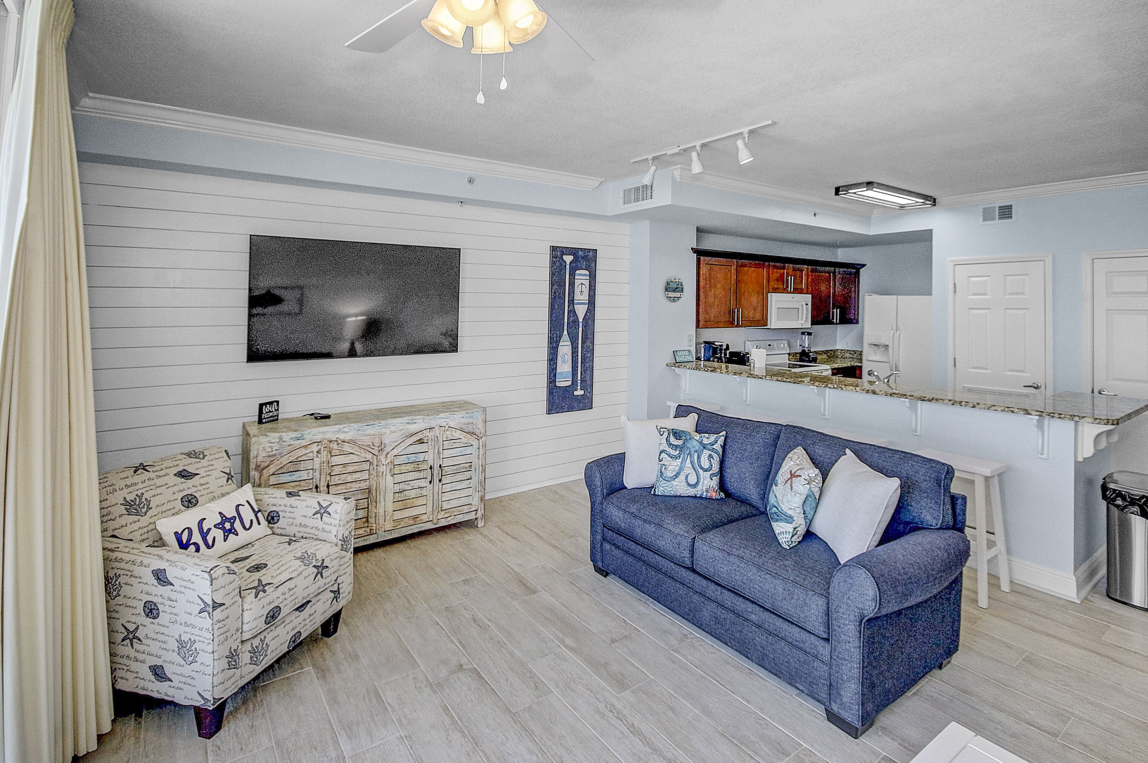 A living room with a blue sofa, a patterned armchair, and a small table. There is a kitchen area with wooden cabinets and a countertop.