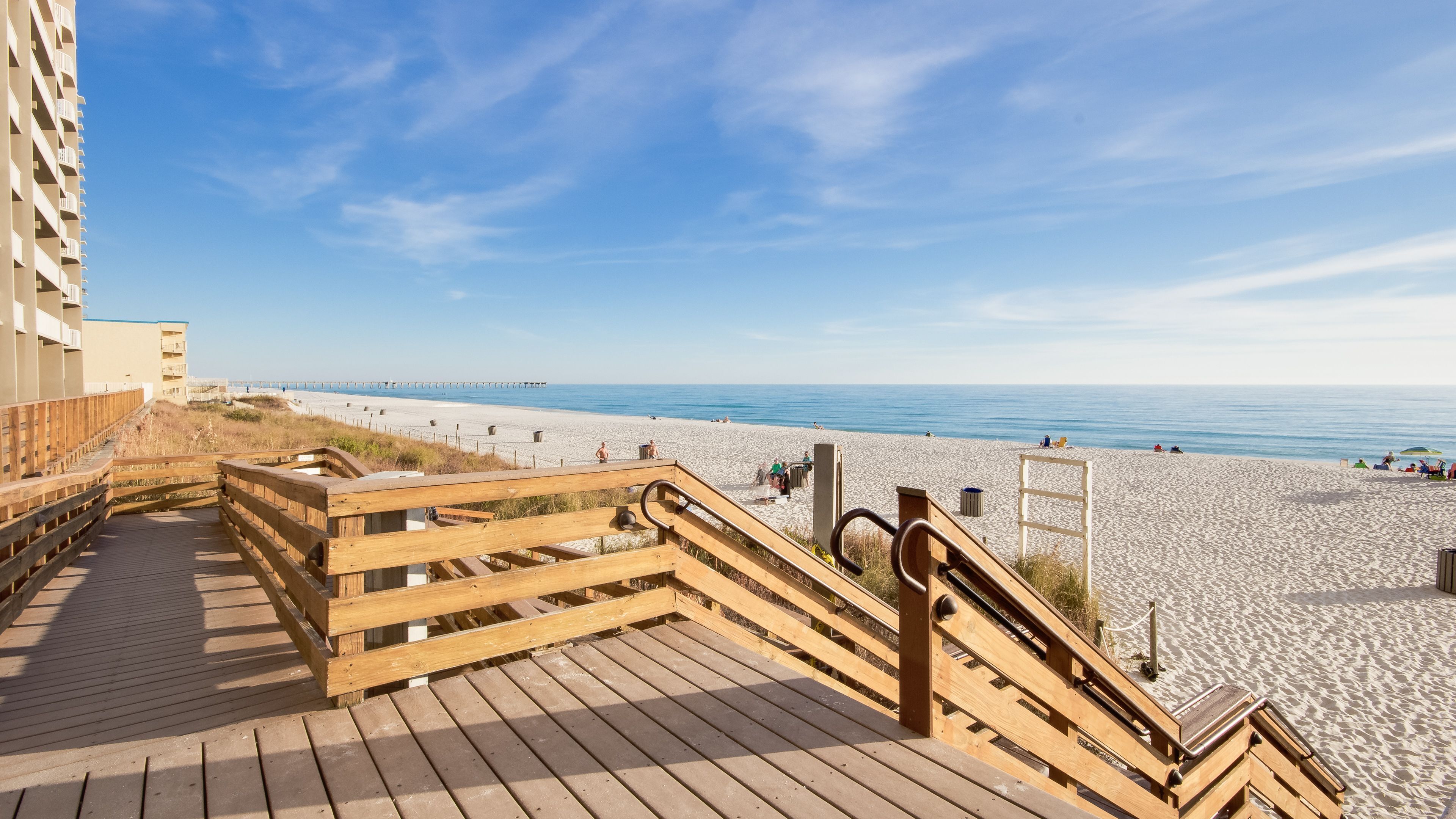 Ein hölzerner Steg führt zu einem Strand mit freiem Blick auf das Meer und einem Pier in der Ferne.