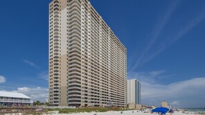 Un hotel alto de varios pisos con numerosas balconadas, situado cerca de una playa con sombrillas y gente disfrutando.