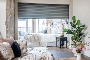 A bedroom with a large bed, a desk, and a chair. There is a window with blinds, a plant, and a vase of flowers.
