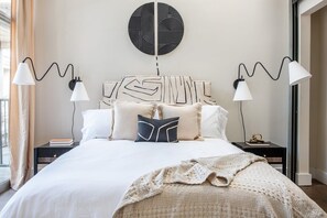 Quarto moderno com uma cama grande, duas mesas de cabeceira, uma obra de arte na parede e uma janela com cortinas.