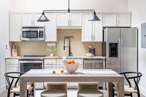 A modern kitchen with white cabinets, a stainless steel refrigerator, a microwave, and a stove. There is a dining table with chairs and a bowl of fruit on the counter.