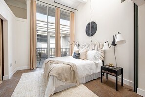 Une chambre avec un grand lit, une table de chevet, une lampe fixée au mur et une vue sur l’extérieur par une fenêtre avec des rideaux.