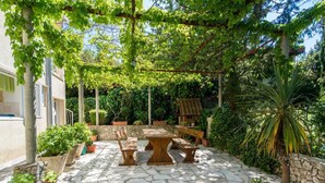 Een terras met stenen vloer, een houten tafel en stoelen, potplanten en een pergola begroeid met klimplanten.