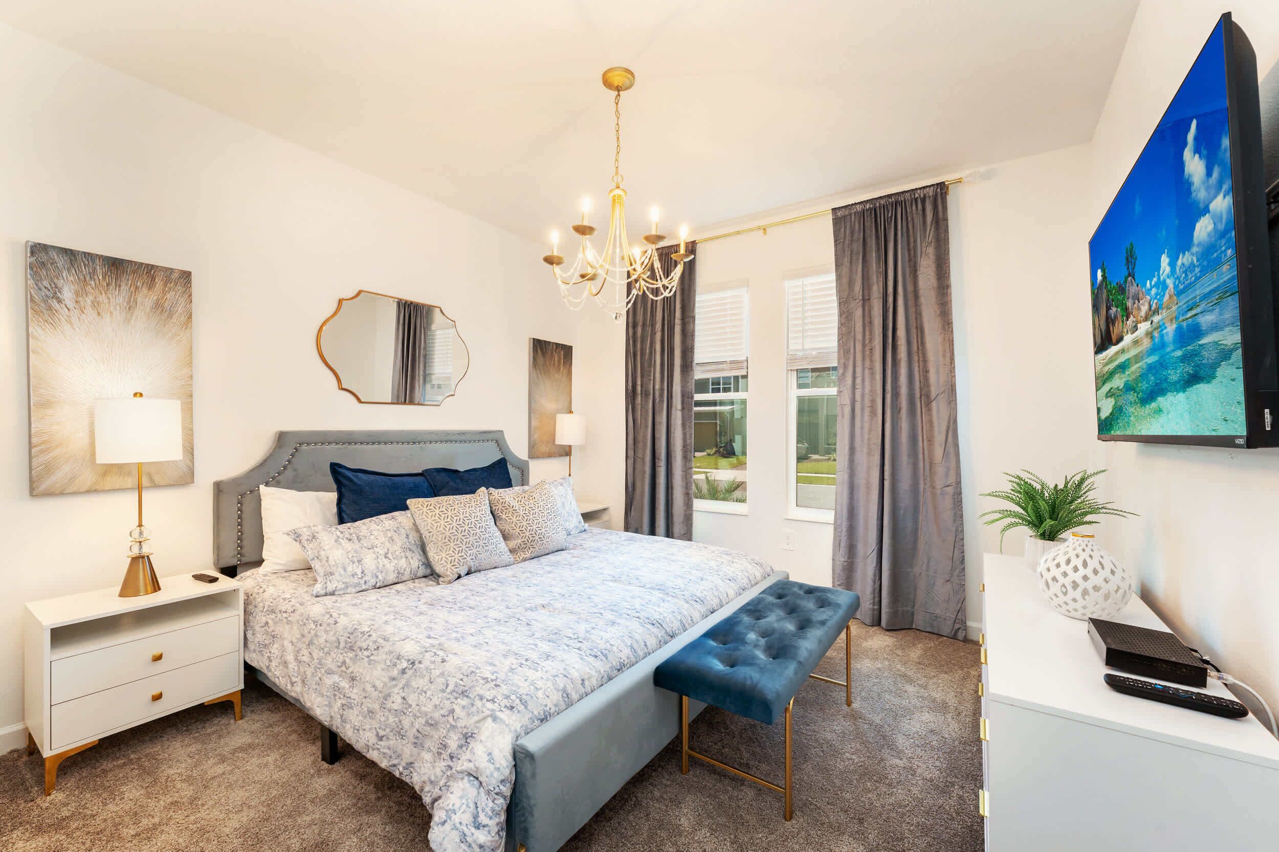 A modern bedroom with a large bed, bedside tables, a chandelier, a painting, and a TV.