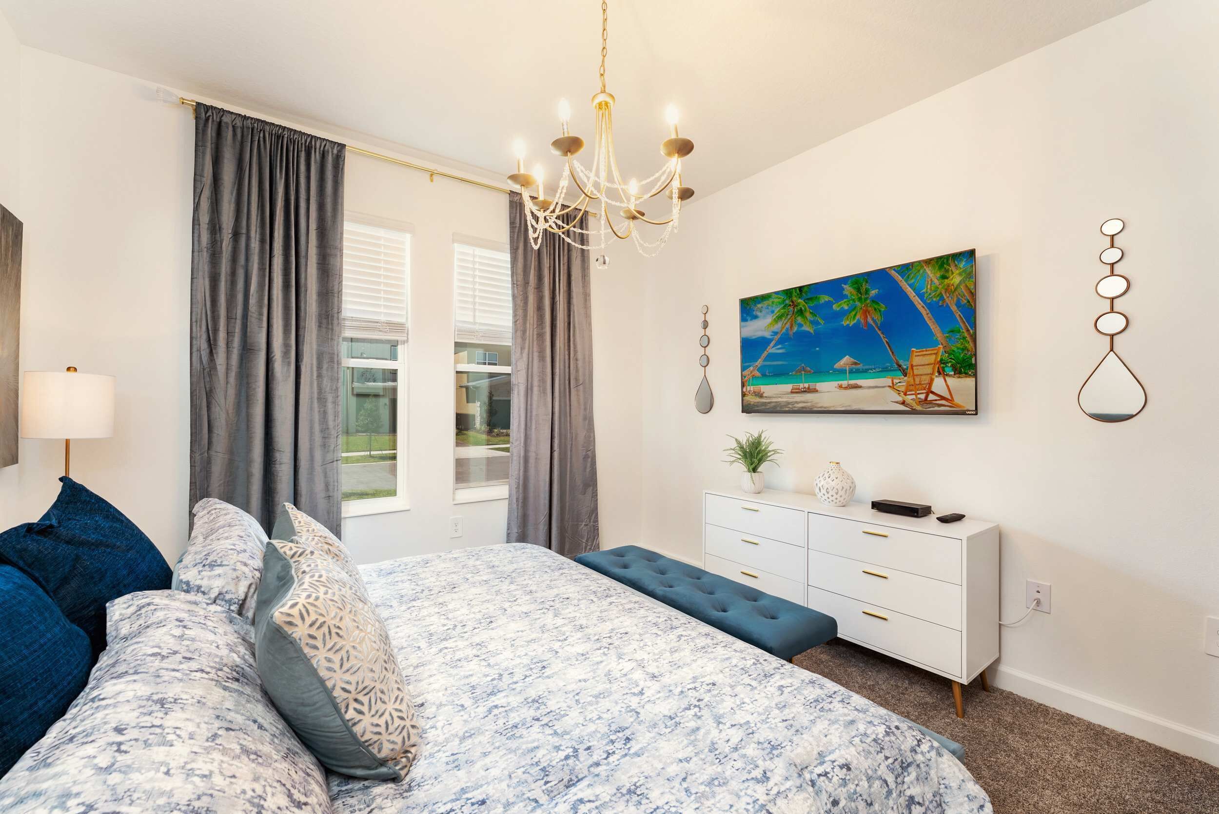 A bedroom with a bed, a dresser, a bench, a chandelier, a painting of a beach, and a window with curtains.