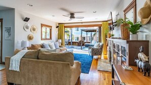 A spacious living area with a beige sofa, a wooden coffee table, and a dining table. There is a large window with curtains, a ceiling fan, and a view of the outdoors. - Mission Beach Retreat, Steps to Bay & Beach (San Diego)