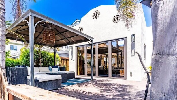 A modern outdoor patio with a covered seating area, a glass-enclosed room, and a white building with circular windows. - Mission Beach Retreat, Steps to Bay & Beach (San Diego)