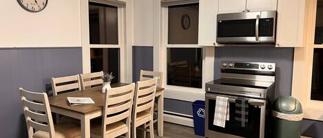 A kitchen with wooden chairs, a dining table, a microwave, an oven, and a clock on the wall.