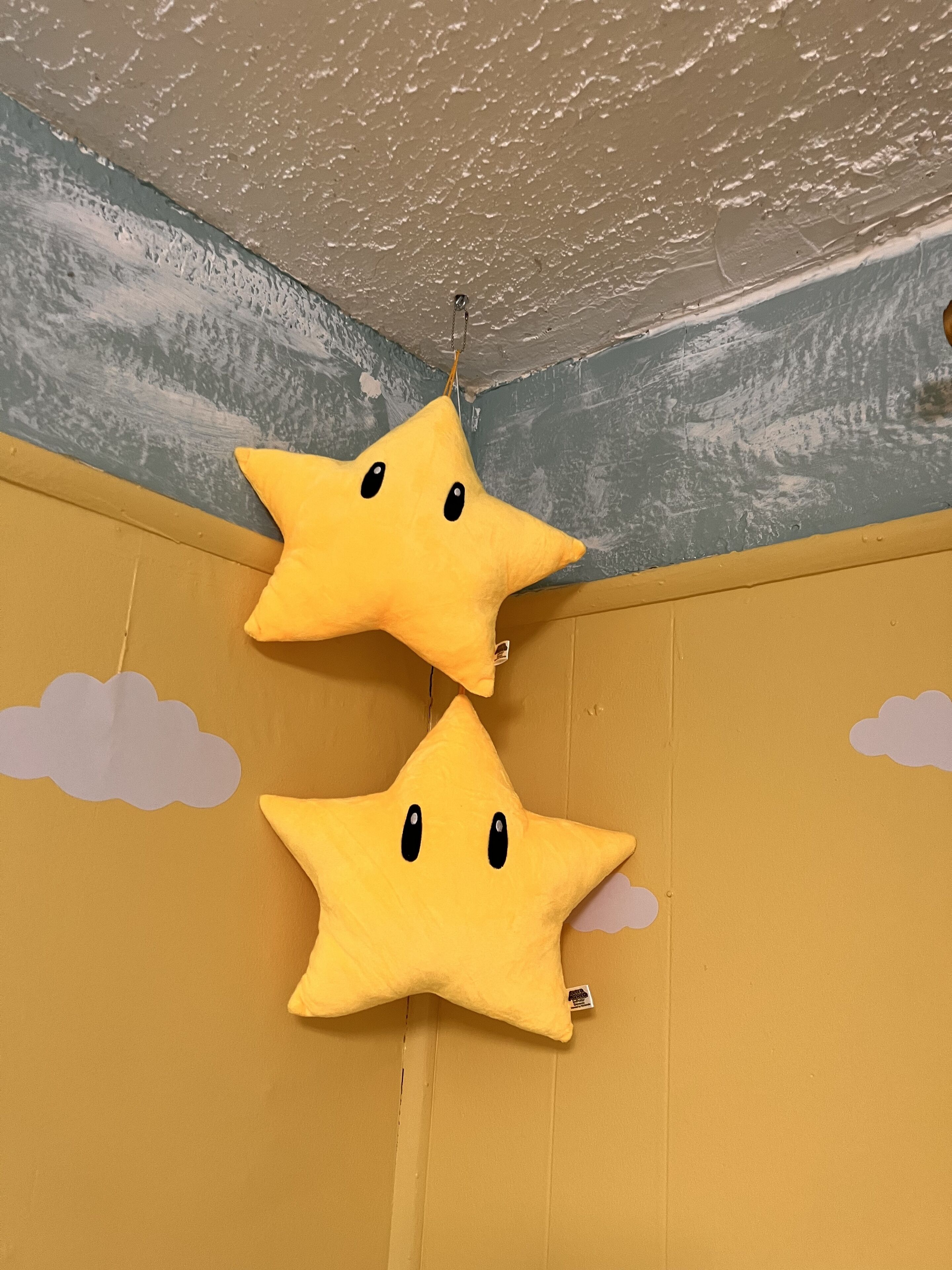 Twee gele ster-vormige kussens hangen aan het plafond in een kamer met een gestructureerde muur en een zichtbare hoek.