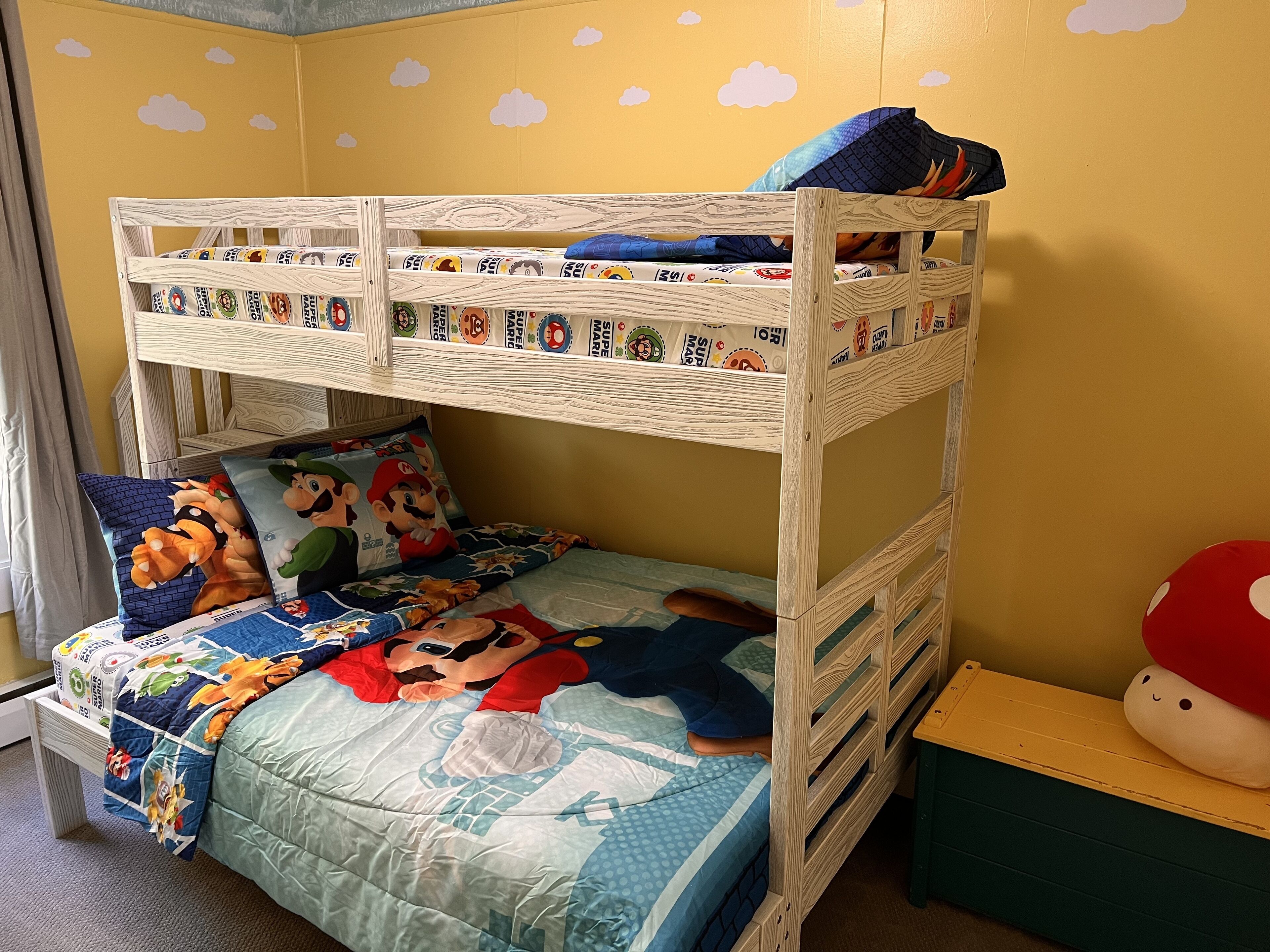 Uma cama beliche com roupa de cama e almofadas com tema do Mario, uma cabeceira de madeira e um criado-mudo pequeno e verde com um brinquedo de pelúcia.