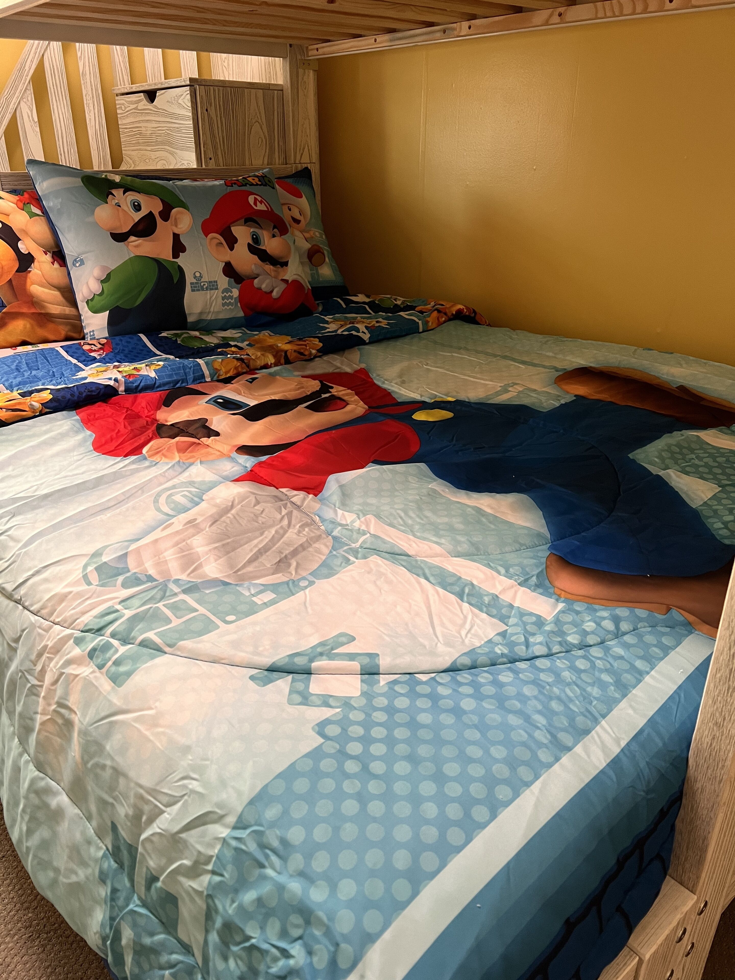 A bedroom with a bed covered in a Mario-themed duvet, a pillow with Mario and Luigi, and a wooden headboard.