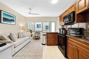 A modern kitchen with wooden cabinets, a microwave, and a stove. There's a sofa and a chair in the living area, and a staircase leading to an upper level.