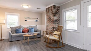 A living room with a gray sofa, a wooden rocking chair, a coffee table, and a brick accent wall. - Beachwalk #4, 8 minute walk to the beach (Virginia Beach)