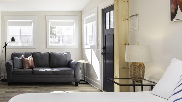 Et moderne hotelværelse med en grå sofa, en seng, et lille bord med en lampe og en dør, der fører til et andet rum.