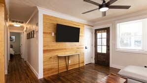 A modern living room with a wooden accent wall, a flat-screen TV, a ceiling fan, and a bench. - Beach Vibe with Hot tub! 10 min walk to the Beach! Amazing Backyard! (Virginia Beach)