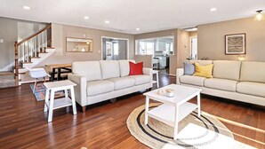 A spacious living room with a wooden staircase, a dining area, and a kitchen visible through an open doorway. - The VIBE House with POOL! (Virginia Beach)