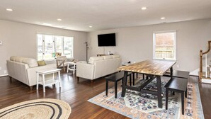 A spacious living area with a wooden dining table, white sofas, and a flat-screen TV mounted on the wall. - The VIBE House with POOL! (Virginia Beach)