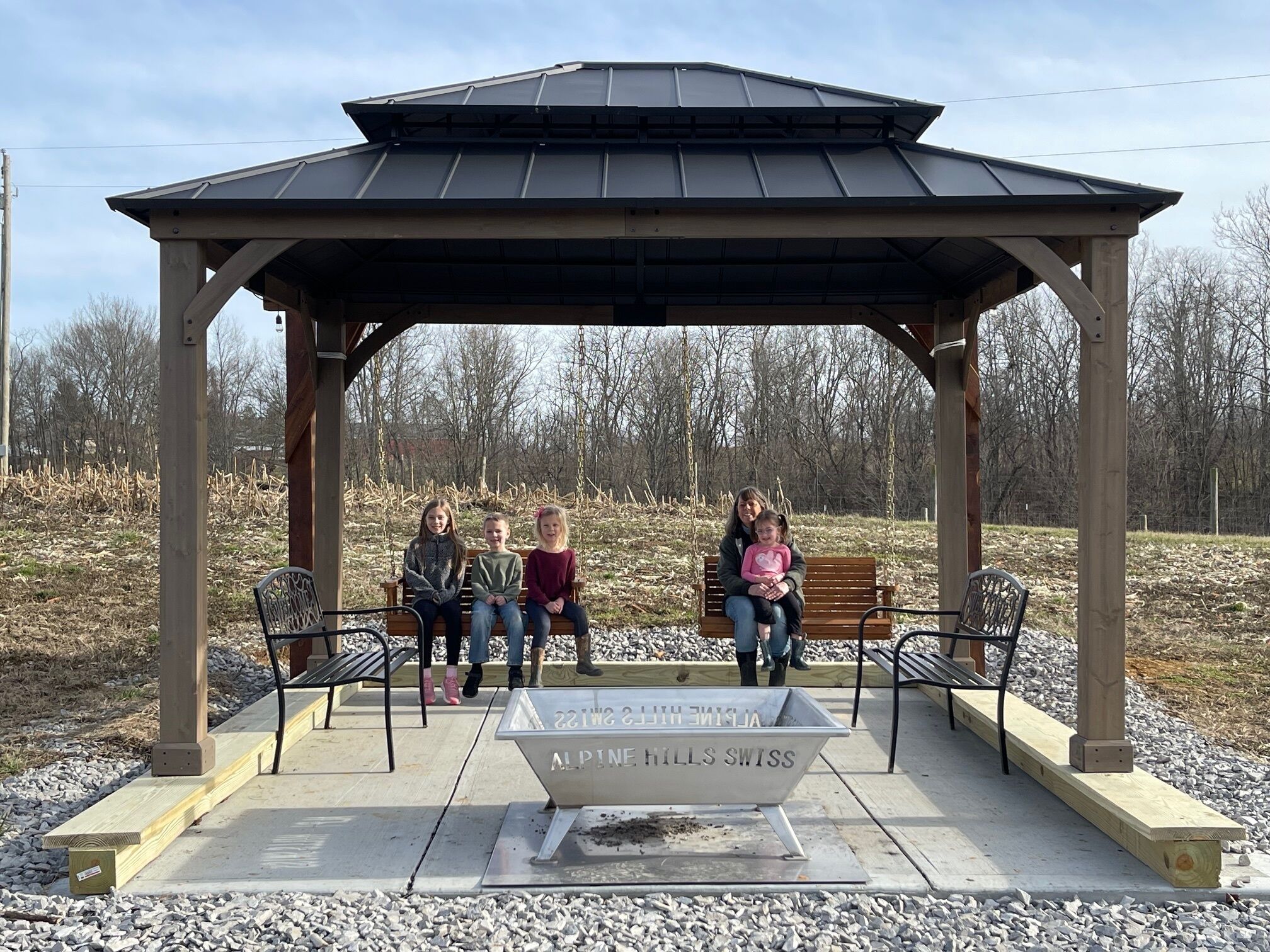 A gazebo with a bench and a metal tub labeled "Alpine Hills Swiss" in a park.