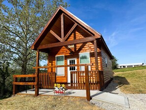 A log cabin with a porch, a welcome sign, and a view of the surrounding landscape.