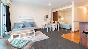 Ein modernes Wohnzimmer mit Sofa, Couchtisch und Essbereich mit Tisch und Stühlen. Im Hintergrund ist ein Bett zu sehen und eine Tür, die in einen anderen Raum führt.