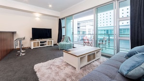 Ein modernes Wohnzimmer mit großem Fernseher, einer Couch, einem Couchtisch und einem Balkon mit Blick auf ein Gebäude.