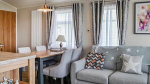 Una habitación acogedora con una mesa de comedor de madera, un sofá gris con cojines de estrellas y estampados geométricos, una lámpara y cortinas grises.