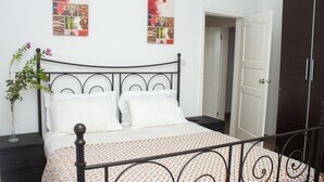 A neatly made bed with a patterned bedspread, a bedside table with a vase of flowers, and a wall with a fruit collage. - Kerr Ayo's One Bedroom Apartment, 5 minutes Away from Senegambia Beaches. (Serrekunda)
