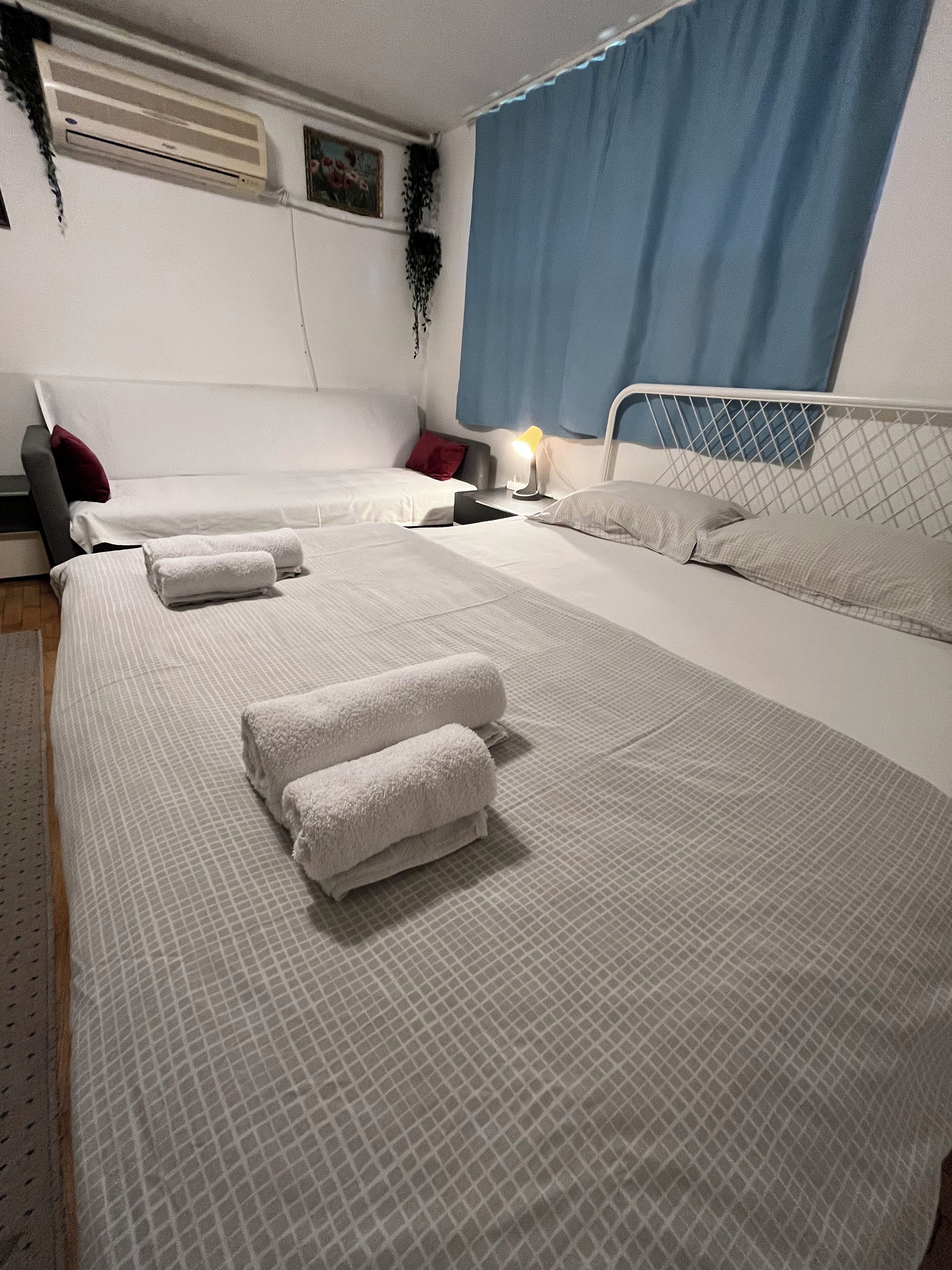A hotel room with a large bed, two bedside tables, a nightstand, an air conditioning unit, and a window with curtains.