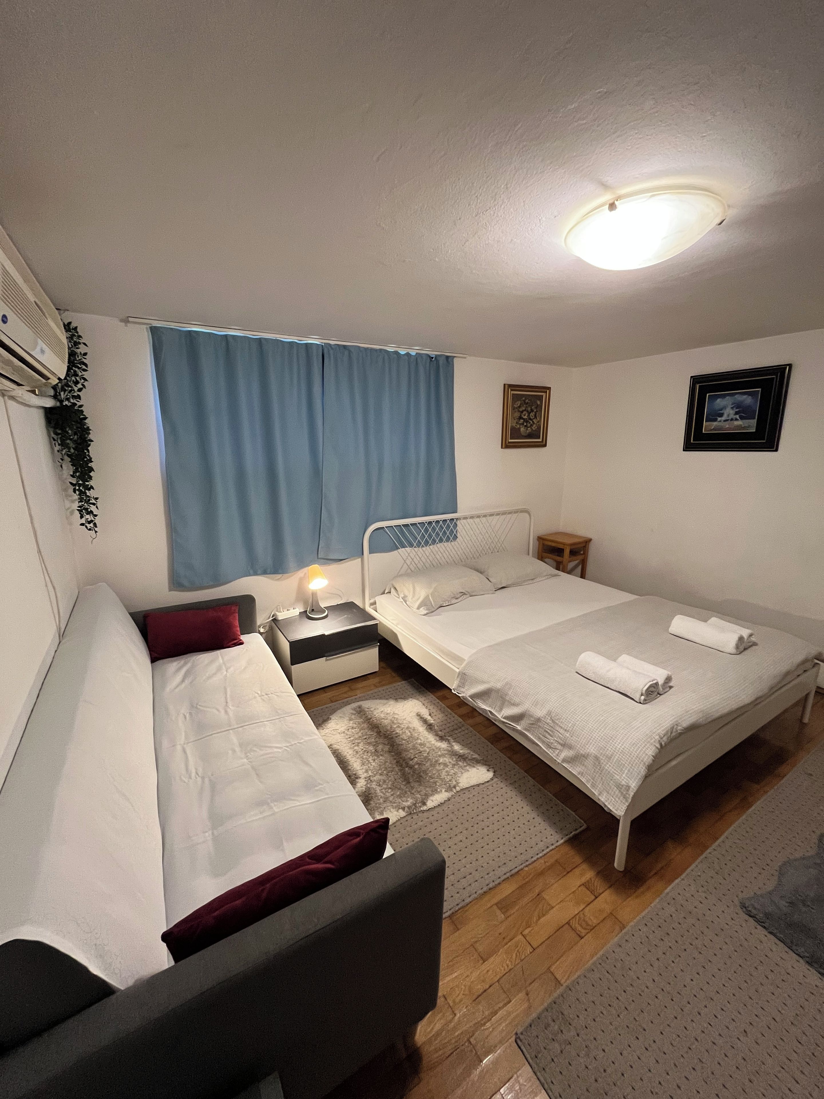 A bedroom with two beds, a nightstand, a lamp, a window with curtains, and a ceiling light.
