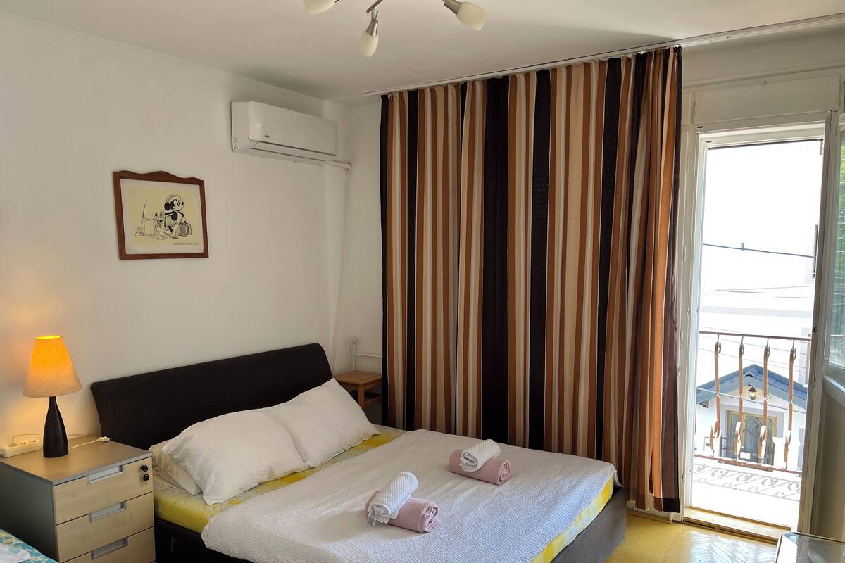 Une chambre d’hôtel comprenant un lit, une lampe de chevet, une petite table, une fenêtre avec des rideaux et un appareil de climatisation.