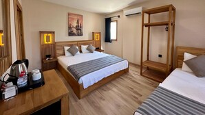 A hotel room with a large bed, a bedside table with a kettle and water bottle, a wooden bench, a wall-mounted air conditioner, and a painting.