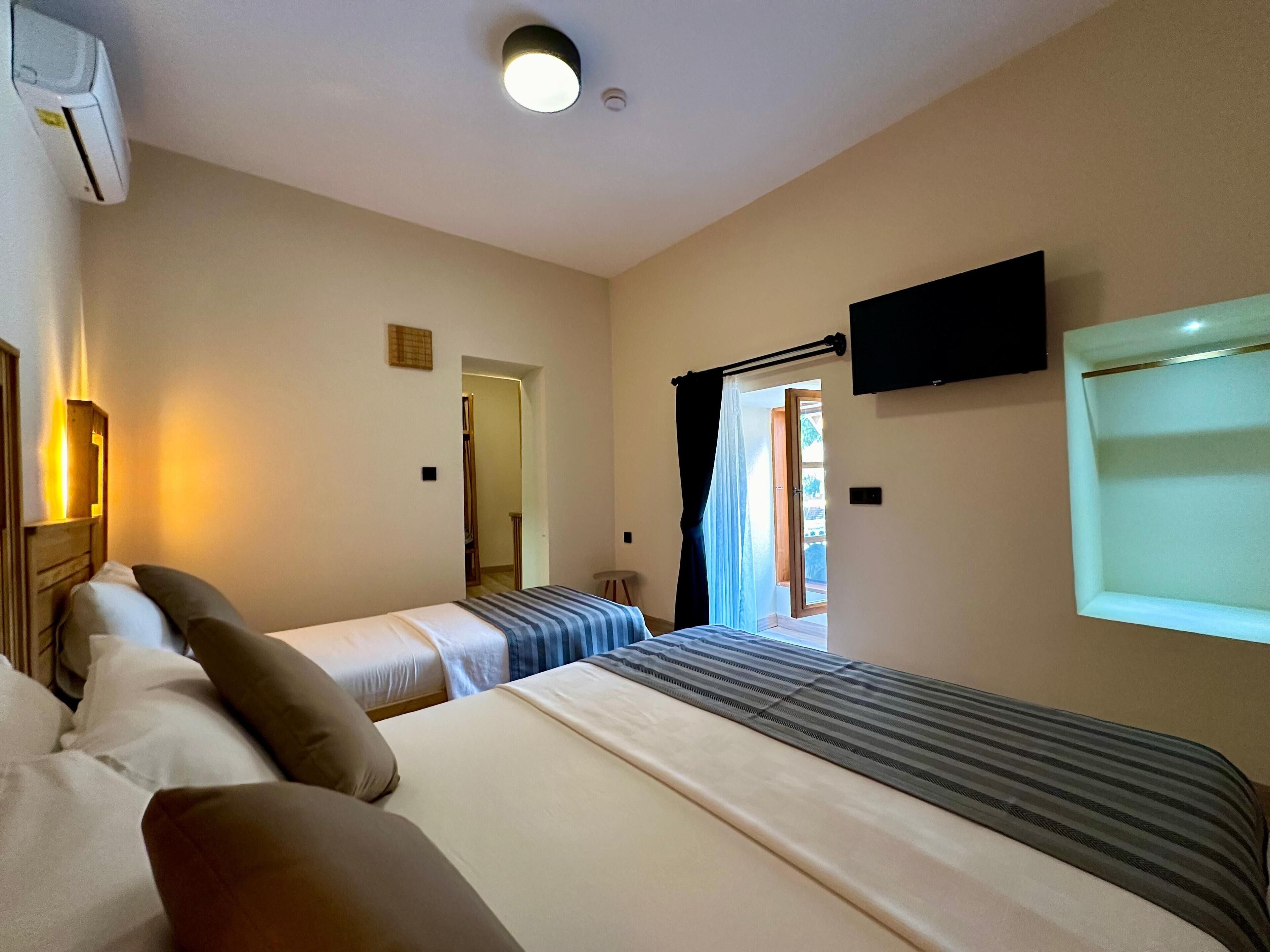 A hotel room with two beds, a flat-screen TV mounted on the wall, a ceiling light, and a window with curtains.