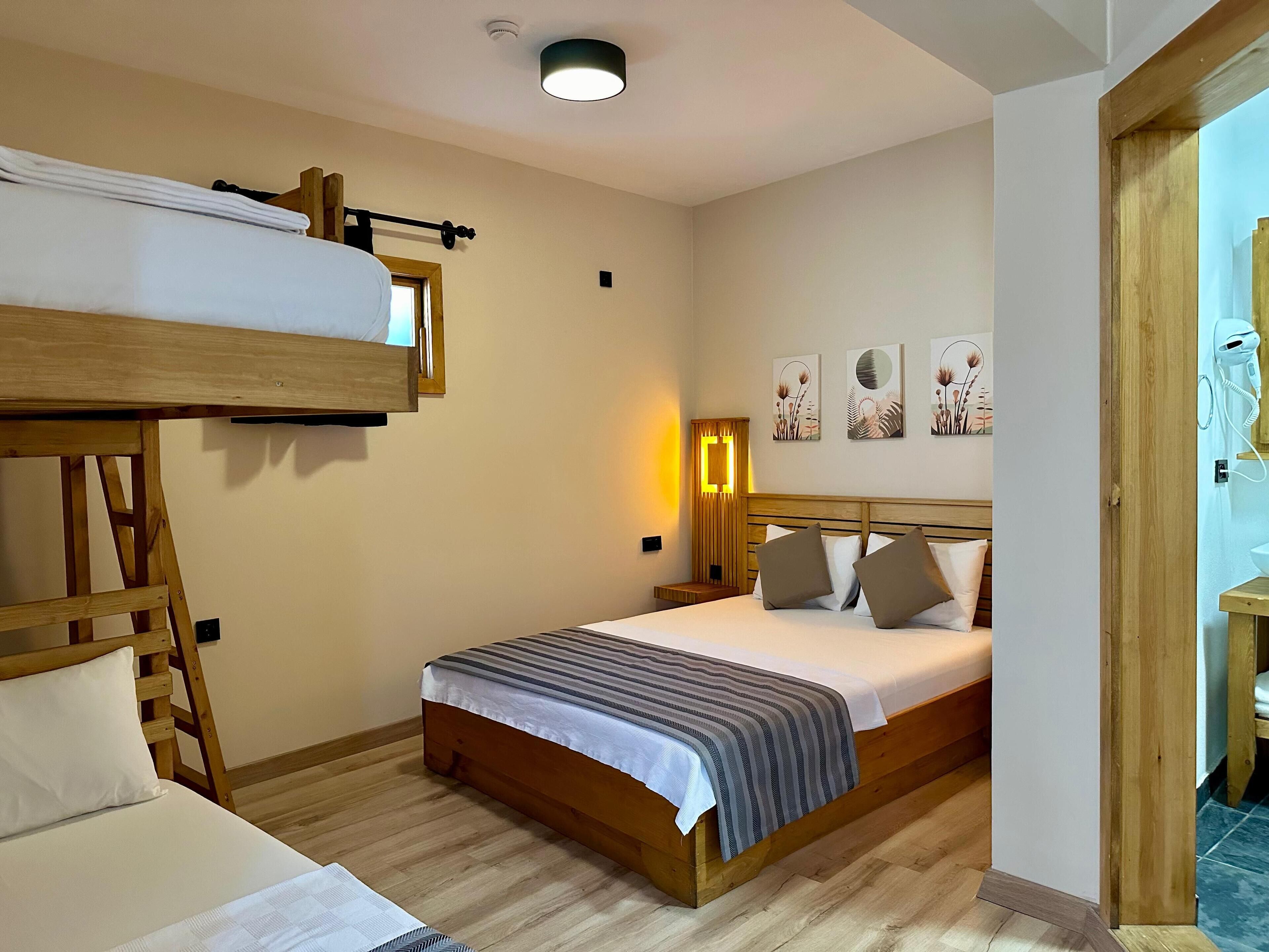 A bedroom with a bunk bed, a single bed, wooden flooring, a bedside table with a lamp, and wall art.