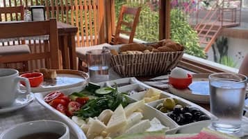 Une table de petit-déjeuner avec divers aliments, un verre d’eau, et en arrière-plan, une vue sur un jardin et des bâtiments.
