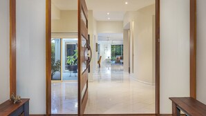 A spacious hallway with a polished floor, wooden benches, and a sliding glass door leading to an outdoor area with plants. - Walk to Golf Course, Lagoon Pool, Coastal Lux Home (Pelican Waters)